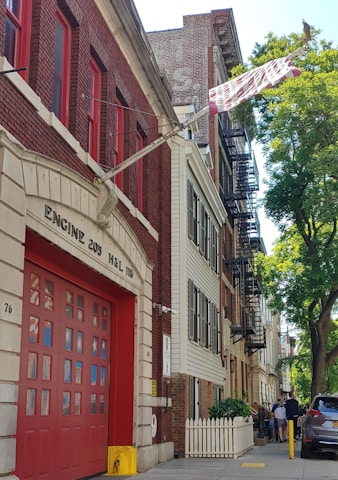 A red brick fire station with large garage doors is positioned on a residential street. The building is labeled 'Engine 205 H&L 118.' Beside it are multi-story residential buildings with fire escapes. A group of people stands on the sidewalk near a white picket fence, and there is a tree providing shade.
