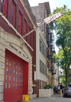 A red brick fire station with large garage doors is positioned on a residential street. The building is labeled 'Engine 205 H&L 118.' Beside it are multi-story residential buildings with fire escapes. A group of people stands on the sidewalk near a white picket fence, and there is a tree providing shade.