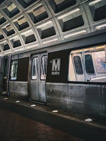 A metro train is stationed at an underground platform with a geometric, concrete ceiling. The train doors are closed, and the lighting gives a slightly moody ambiance. The distinguishing feature is the large 'M metro' logo prominently displayed on the side of the train.