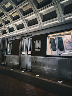 A metro train is stationed at an underground platform with a geometric, concrete ceiling. The train doors are closed, and the lighting gives a slightly moody ambiance. The distinguishing feature is the large 'M metro' logo prominently displayed on the side of the train.