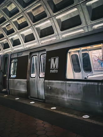 A metro train is stationed at an underground platform with a geometric, concrete ceiling. The train doors are closed, and the lighting gives a slightly moody ambiance. The distinguishing feature is the large 'M metro' logo prominently displayed on the side of the train.