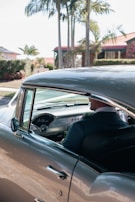 A vintage car with a well-maintained classic interior is parked under palm trees. The driver, dressed in a formal suit, is visible through the open window.