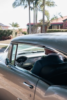A vintage car with a well-maintained classic interior is parked under palm trees. The driver, dressed in a formal suit, is visible through the open window.
