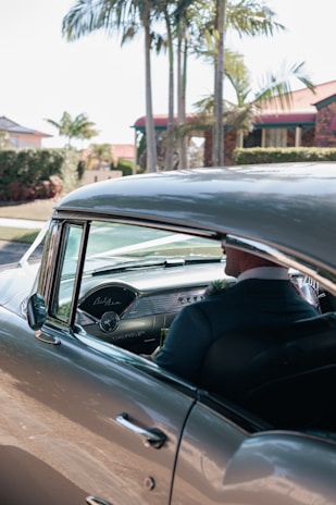 A vintage car with a well-maintained classic interior is parked under palm trees. The driver, dressed in a formal suit, is visible through the open window.