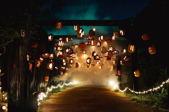 An atmospheric scene with numerous carved pumpkins, each illuminated to create a glowing effect, are suspended in mid-air over a path. A hazy fog adds to the mysterious ambiance, while string lights line the walkway, providing a soft, warm glow. The surrounding trees and dark night sky enhance the magical Halloween setting.