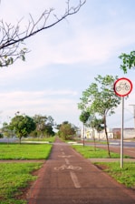 A paved pathway marked for bicycles stretches into the distance, flanked by grassy areas and trees on both sides. A bicycle sign is prominently displayed, indicating that the path is meant for cycling. The sky is clear with few clouds, suggesting a pleasant day.