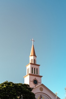 A church steeple with a cross on top, set against a clear blue sky. The building is made of a light-colored material, and there is a large circular window. The bottom part of the image shows some leafy green trees.