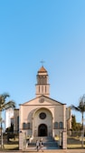 A church with a traditional architectural design featuring a cross at the top of its steeple, positioned under a clear blue sky. The building is framed by palm trees on either side, and a couple of people can be seen walking in front of the church.