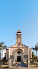 A church with a traditional architectural design featuring a cross at the top of its steeple, positioned under a clear blue sky. The building is framed by palm trees on either side, and a couple of people can be seen walking in front of the church.