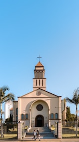 A church with a traditional architectural design featuring a cross at the top of its steeple, positioned under a clear blue sky. The building is framed by palm trees on either side, and a couple of people can be seen walking in front of the church.
