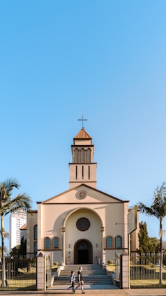 A church with a traditional architectural design featuring a cross at the top of its steeple, positioned under a clear blue sky. The building is framed by palm trees on either side, and a couple of people can be seen walking in front of the church.