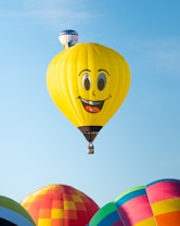 A bright yellow hot air balloon with a large smiling face is prominently featured in the sky, surrounded by other colorful balloons. The scene conveys a vibrant and cheerful atmosphere, with clear blue skies providing a picturesque backdrop.