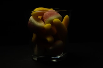 A glass filled with colorful gummy candies set against a dark background. The candies are mostly yellow and pinkish-white in color, with a sugary texture that catches the light.