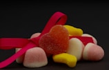 Several colorful sugar-coated gummy candies are arranged with a bright pink ribbon. A heart-shaped red-orange candy is prominently placed in the center, surrounded by pink and white cone-shaped candies and yellow banana-shaped candies. The dark background enhances the vivid colors of the candies and ribbon.