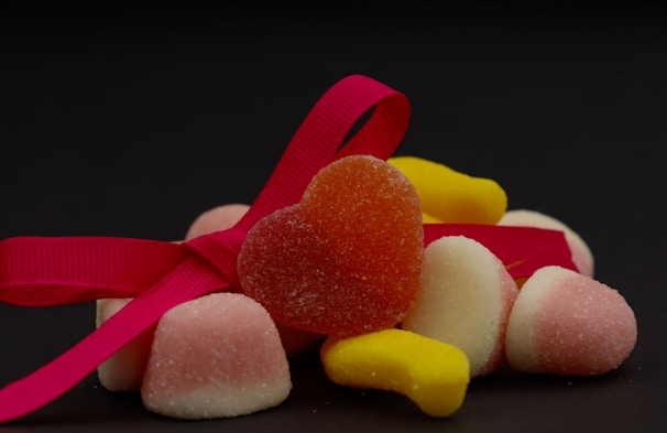 Several colorful sugar-coated gummy candies are arranged with a bright pink ribbon. A heart-shaped red-orange candy is prominently placed in the center, surrounded by pink and white cone-shaped candies and yellow banana-shaped candies. The dark background enhances the vivid colors of the candies and ribbon.
