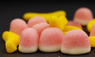 A pile of colorful gummy candies, including pink and white dome-shaped ones and elongated yellow ones, covered in a sugary coating, set against a dark background.