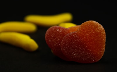 A close-up view of heart-shaped orange gummies dusted with sugar in the foreground. In the background, there are several banana-shaped yellow gummies.