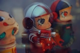 Three vinyl toy figures with oversized heads and exaggerated eyes. The central figure is dressed in a space suit, holding a red canister with a Coca-Cola logo. The figure on the right wears a green outfit with a red and black helmet. The figure on the left is partially visible with a smiling expression.
