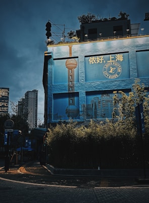 A building facade displays a large advertisement for a Louis Vuitton City Guide featuring the skyline of Shanghai, with the Oriental Pearl Tower prominently featured. The scene is illuminated by dim streetlights, creating a moody urban evening atmosphere. Surrounding the bottom of the building are dense green plants, and the sky above is a deep blue, suggesting either dawn or dusk.