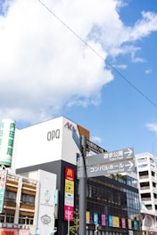 A street scene featuring a large commercial building with various signs, including a McDonald's logo and directions to Yuho Park and Compal Hall. The sky is clear with large, fluffy clouds, and there are other buildings in the background.