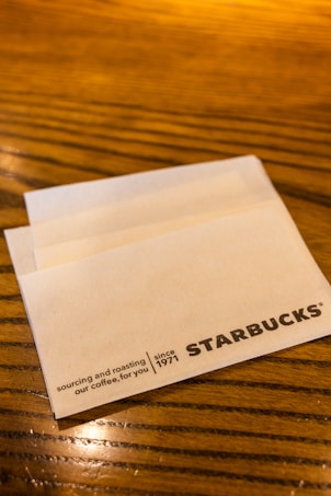 A wooden table surface with a white napkin featuring the Starbucks logo and text indicating the company's focus on sourcing and roasting coffee since 1971.