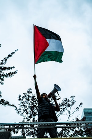 A person stands on an elevated platform holding a megaphone and raising a flag with black, white, and green horizontal stripes and a red triangle. The backdrop includes trees and a partly cloudy sky.