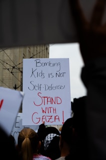 A protest sign held by a crowd contains the message 'Bombing Kids is Not Self-Defense, Stand with Gaza!' with people gathered around, some wearing headscarves.