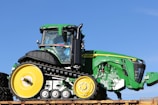 A large, green vehicle with black tracks and yellow wheels is prominently displayed against a clear blue sky. The vehicle features a modern, angular design with visible branding and a well-defined cab area.