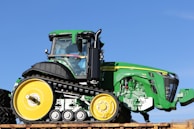 A large, green vehicle with black tracks and yellow wheels is prominently displayed against a clear blue sky. The vehicle features a modern, angular design with visible branding and a well-defined cab area.