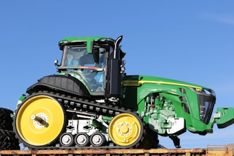 A large, green vehicle with black tracks and yellow wheels is prominently displayed against a clear blue sky. The vehicle features a modern, angular design with visible branding and a well-defined cab area.