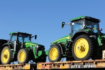 Two green tractors with yellow wheels are positioned on a flatbed rail car. The tractors have a modern design and are likely intended for agricultural use. The clear blue sky serves as a backdrop, enhancing the vivid colors of the machinery.