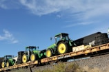Several green and yellow tractors are loaded onto a long flatbed railcar. The sky is clear and blue with a few clouds. The railcar is on a track with some vegetation on the surrounding ground.