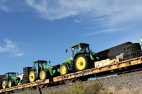 Several green and yellow tractors are loaded onto a long flatbed railcar. The sky is clear and blue with a few clouds. The railcar is on a track with some vegetation on the surrounding ground.