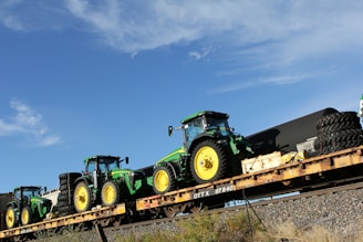 Several green and yellow tractors are loaded onto a long flatbed railcar. The sky is clear and blue with a few clouds. The railcar is on a track with some vegetation on the surrounding ground.