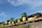Several green and yellow tractors are loaded onto a long flatbed railcar. The sky is clear and blue with a few clouds. The railcar is on a track with some vegetation on the surrounding ground.
