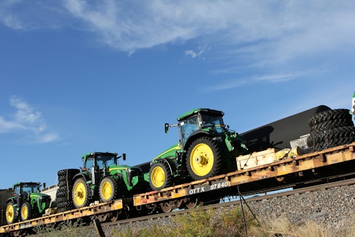 Several green and yellow tractors are loaded onto a long flatbed railcar. The sky is clear and blue with a few clouds. The railcar is on a track with some vegetation on the surrounding ground.