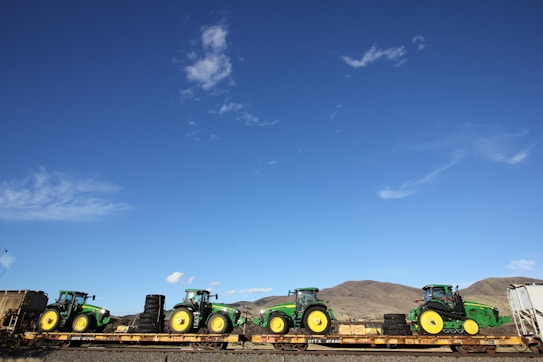 A series of green tractors are secured on a flatbed train car. They are in a rural setting with mountains in the background and a clear blue sky overhead. The tractors' bright yellow wheels contrast with the sunny landscape.