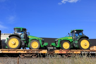 Two green tractors with large yellow wheels are placed on flatbed train cars. The tractors appear new and display branding. The sky is clear and blue in the background.