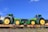 Two green tractors with large yellow wheels are placed on flatbed train cars. The tractors appear new and display branding. The sky is clear and blue in the background.