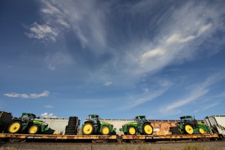 Multiple green tractors are loaded onto a flatbed train car under a bright blue sky with scattered clouds.