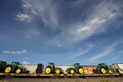 Multiple green tractors are loaded onto a flatbed train car under a bright blue sky with scattered clouds.