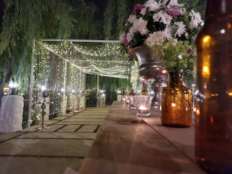 A beautifully decorated outdoor walkway is lined with hanging fairy lights. Along the table, brown glass bottles and small candles create a warm, inviting atmosphere. A large bouquet of white and purple flowers sits in an elegant silver vase to the right. The scene is surrounded by lush greenery, adding to the intimate and festive setting.