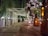 A beautifully decorated outdoor walkway is lined with hanging fairy lights. Along the table, brown glass bottles and small candles create a warm, inviting atmosphere. A large bouquet of white and purple flowers sits in an elegant silver vase to the right. The scene is surrounded by lush greenery, adding to the intimate and festive setting.