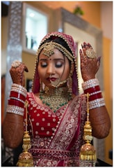 A woman dressed in traditional Indian bridal attire, adorned with intricate jewelry including a maang tikka, necklace, and bangles. Her hands display detailed henna designs, and she wears an ornate red and gold sari. The background features a softly blurred room interior.