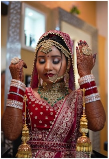 A woman dressed in traditional Indian bridal attire, adorned with intricate jewelry including a maang tikka, necklace, and bangles. Her hands display detailed henna designs, and she wears an ornate red and gold sari. The background features a softly blurred room interior.