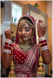 A woman dressed in traditional Indian bridal attire, adorned with intricate jewelry including a maang tikka, necklace, and bangles. Her hands display detailed henna designs, and she wears an ornate red and gold sari. The background features a softly blurred room interior.