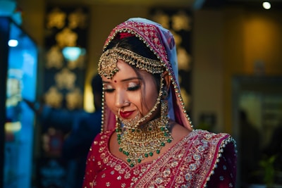 A woman dressed in traditional bridal attire, adorned with intricate gold jewelry, a nose ring, and a vibrant red and pink embellished outfit with a matching headscarf. Her eyes are closed, and she appears to be in a serene and reflective mood.