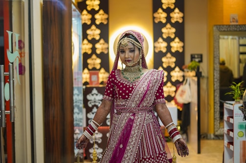 A woman dressed in elaborate traditional attire stands indoors. She wears a beautifully embroidered pink and gold lehenga, with intricate jewelry adorning her head, neck, and wrists. Her hands are decorated with mehndi designs. The background features ornate black and gold decor with soft lighting accentuating the festive atmosphere. Various items are visible behind her, adding to the setting's richness.