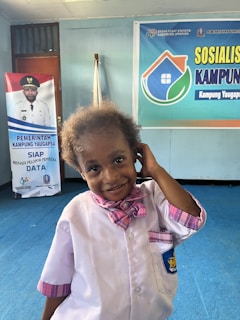 A young child wearing a light pink shirt with matching pink bow tie stands indoors. The child has a playful expression and is touching their ear with one hand. Behind the child, there is a blue banner with colorful text and a logo resembling a house. There's also a standee featuring a person in uniform.