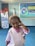 A young child wearing a light pink shirt with matching pink bow tie stands indoors. The child has a playful expression and is touching their ear with one hand. Behind the child, there is a blue banner with colorful text and a logo resembling a house. There's also a standee featuring a person in uniform.